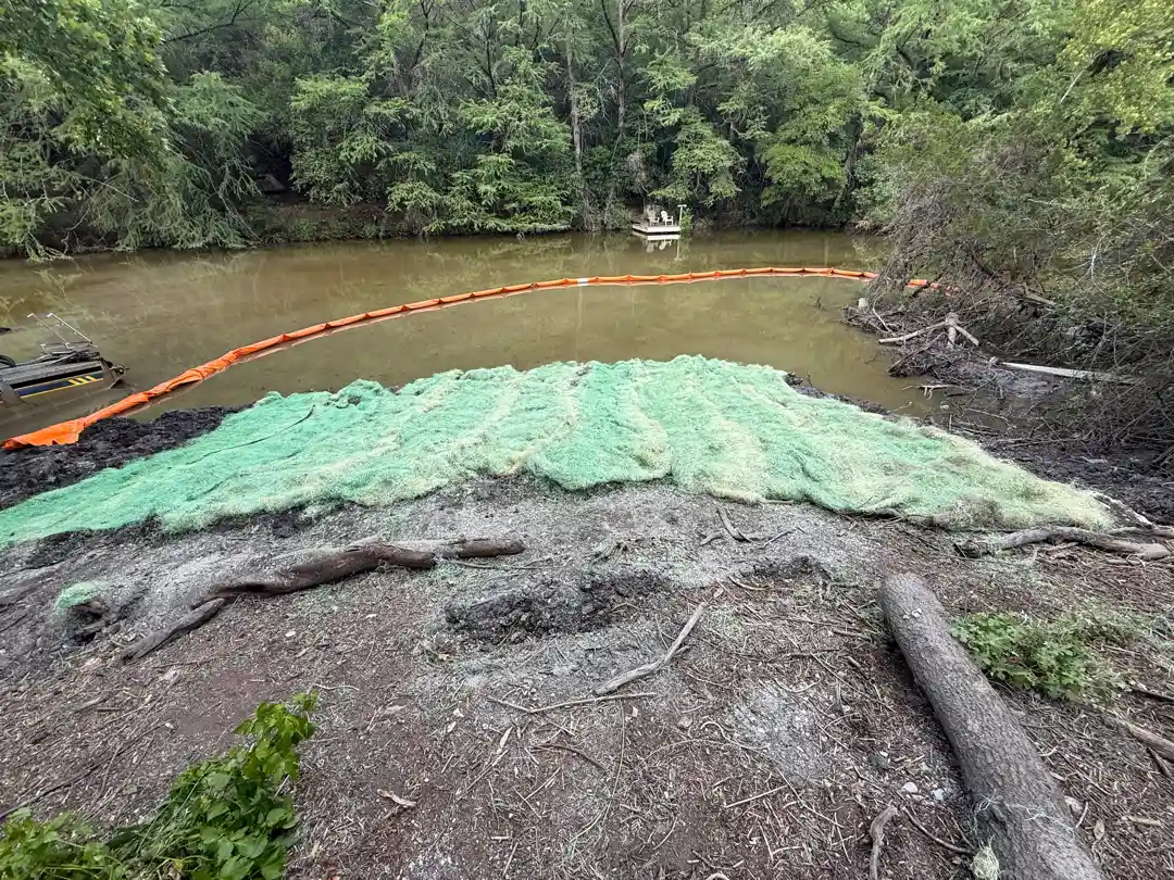 Graded shoreline covered with green erosion-control mat beside a floating orange turbidity curtain.